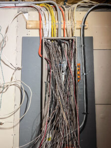 A tangle of various colored cables and wires extending from a panel on a wall, secured with wooden brackets. The wires appear disorganized, with some connections leading to different areas outside the panel.