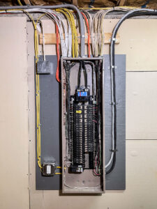 An open electrical panel on a wall, showing multiple circuit breakers and colorful wires neatly organized. The panel is mounted on a board, with cables running through the top and bottom.