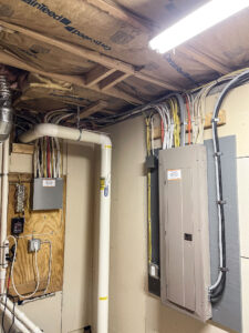 A basement with exposed insulation and wiring on the ceiling. Two electrical panels are mounted on the wall beside a white pipe. Additional wires and cables run along the walls and ceiling, with a few outlets and switches visible.