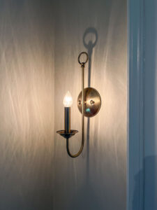 A vintage-style brass wall sconce with a single exposed bulb casts a warm glow, creating intricate shadows on the wall. The design features a circular backplate and a curved arm.