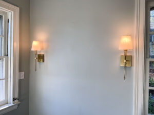 Two wall sconces with white lampshades are mounted on a light gray wall. Each light fixture has a brass finish. Natural light enters through nearby windows, illuminating the room softly.